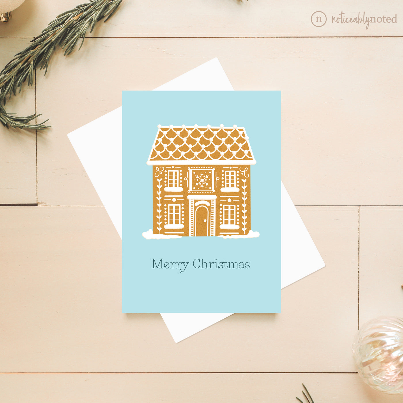 Greeting card with gingerbread house design and 'Merry Christmas' text on a light blue background, placed on a wooden surface.