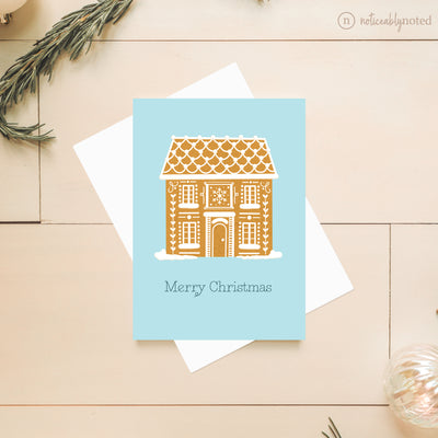 Greeting card with gingerbread house design and 'Merry Christmas' text on a light blue background, placed on a wooden surface.