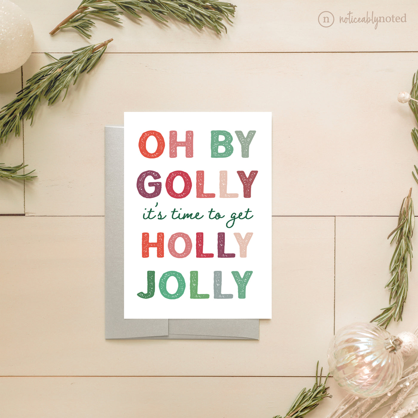 Christmas card with colorful text 'OH BY GOLLY it's time to get HOLLY JOLLY' on a wooden surface with decorative elements.