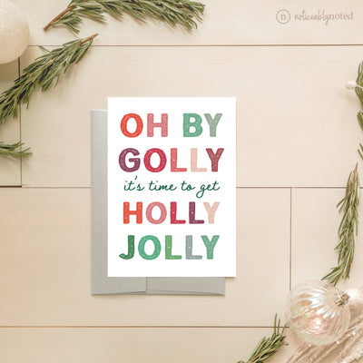 Christmas card with colorful text 'OH BY GOLLY it's time to get HOLLY JOLLY' on a wooden surface with decorative elements.